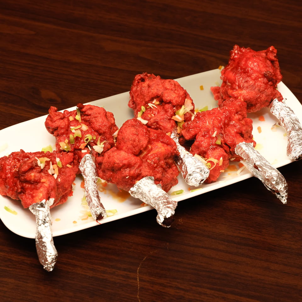 Platter of red-spiced fried chicken drumsticks garnished with chopped vegetables and wrapped foil handles on a white plate.