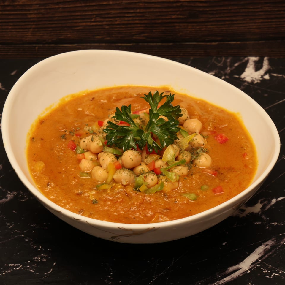 Creamy chickpea curry topped with parsley and diced peppers, served in a white bowl on a black marble table.