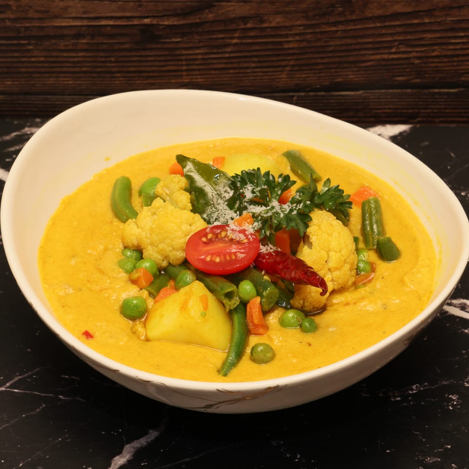 Bowl of creamy yellow vegetable curry topped with cauliflower, green beans, peas, tomato, herbs, and dried chili.