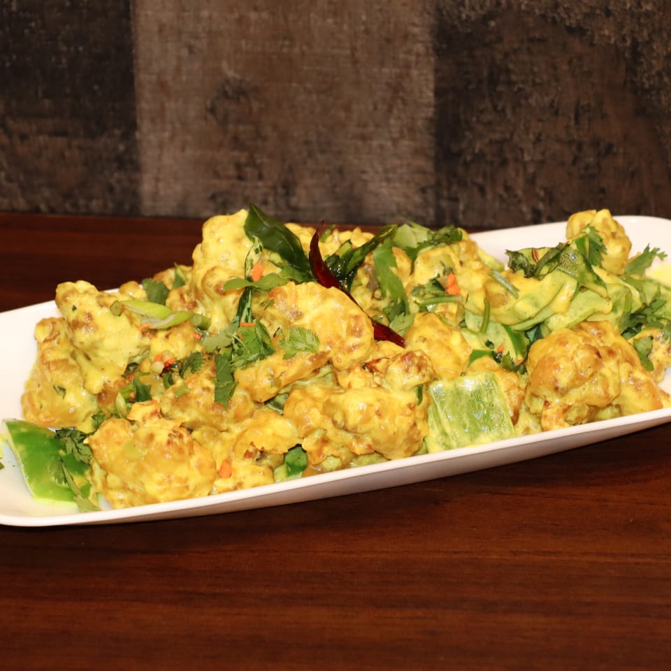 Golden battered cauliflower topped with cilantro, chilies, and vegetables on a white platter.