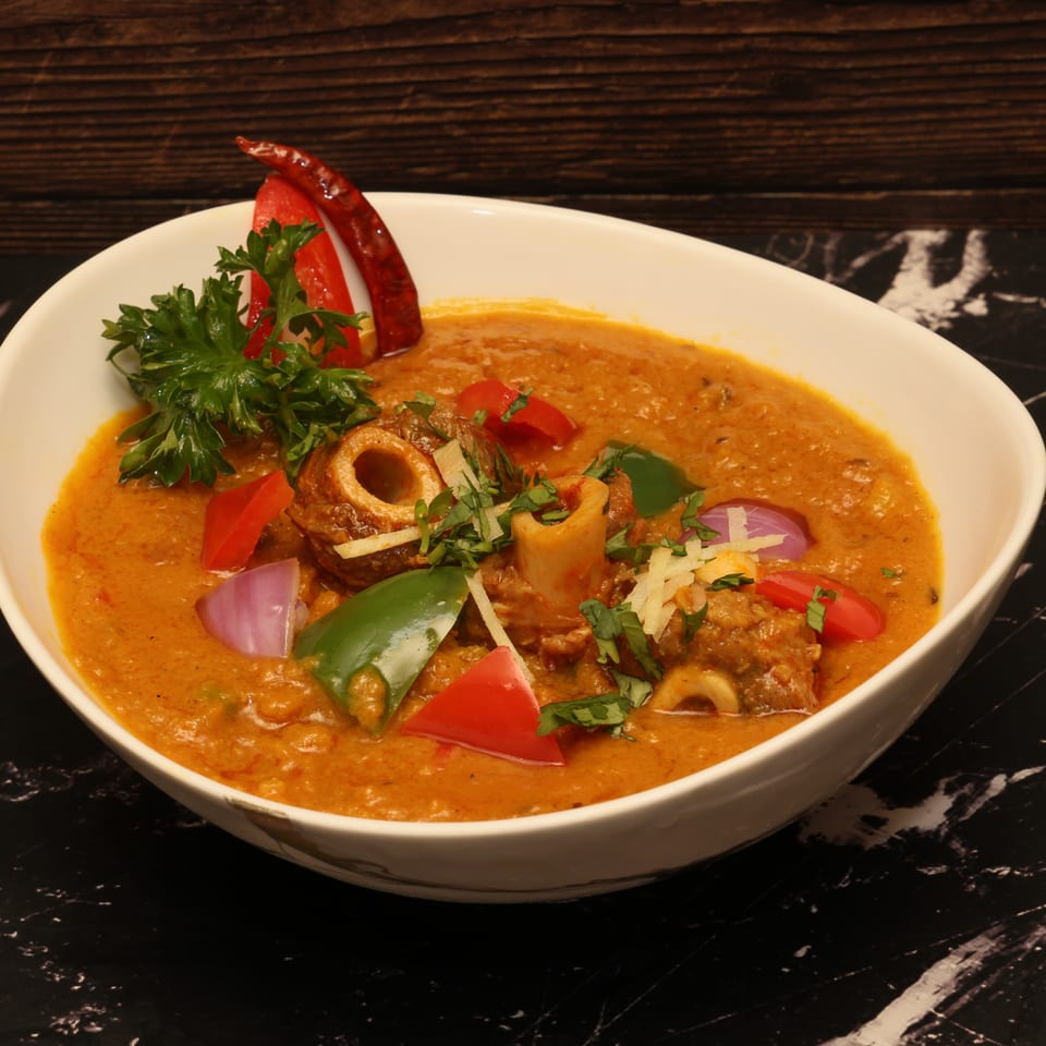Bowl of creamy orange curry with meat, peppers, onions, herbs, ginger, and dried chilies on a black marble table.